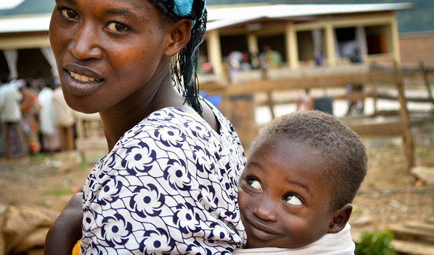 A mom carries her young son in a wrap attached to her back, and he looks up at her admiringly.