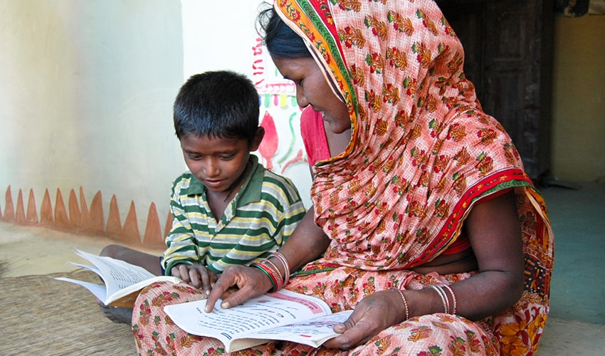 A Nepalese mother dressed in a sari, sits on the floor with her son while they read school books together.