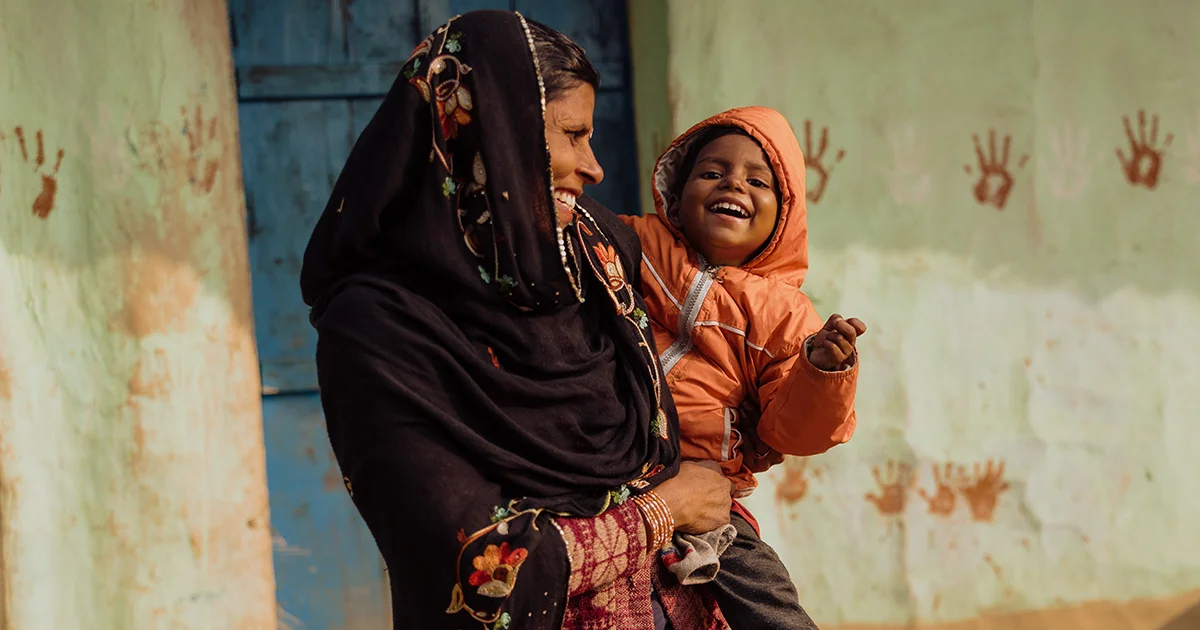A woman holds a small child outside a home with painted handprints on the wall behind them.