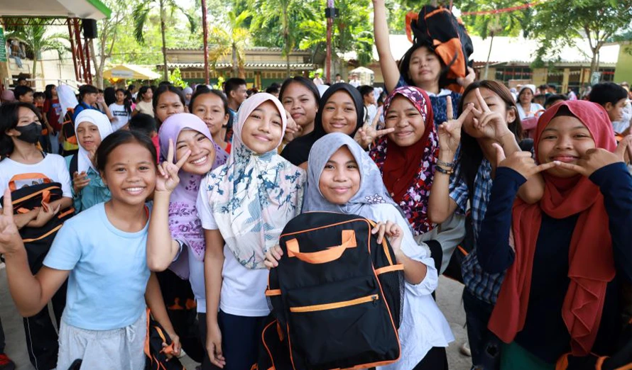 A group of students smiling and holding backpacks full of supplies like notebooks, pens and colouring materials.