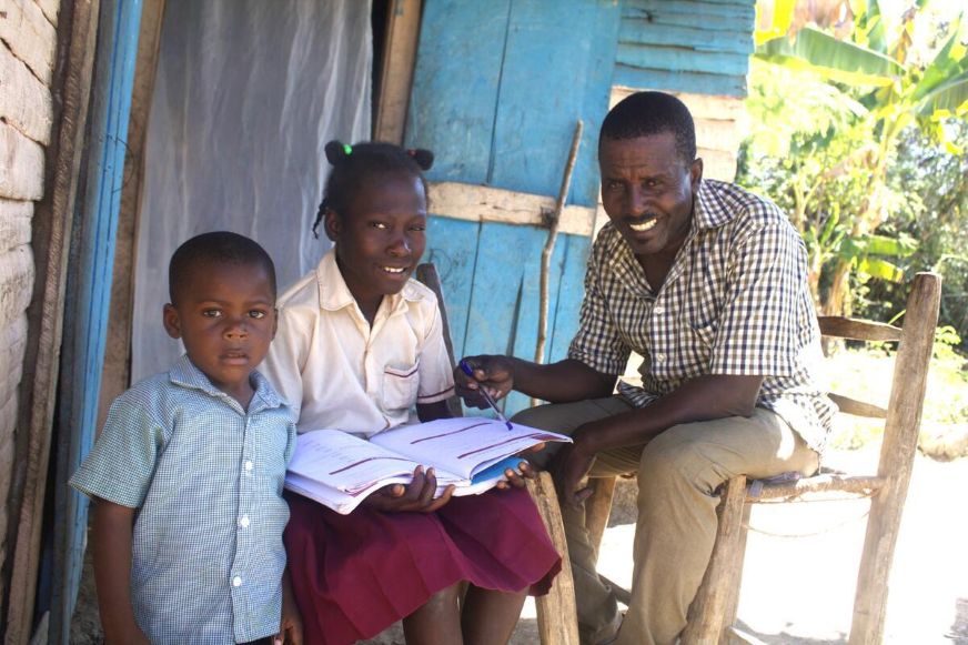 An adult seated outside a small home helps a school-aged child with an open notebook while a younger child stands nearby. (Haiti, 2024)