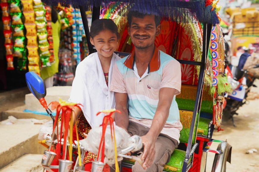 An adult rides a brightly decorated rickshaw with a smiling child seated behind in a busy street setting. (Bangladesh, 2025)