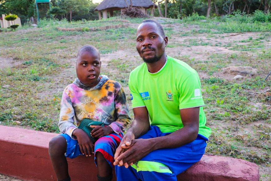 An adult and a child sit side by side outdoors on a low wall, with a small rural home and fields in the background. (Zimbabwe, 2026)