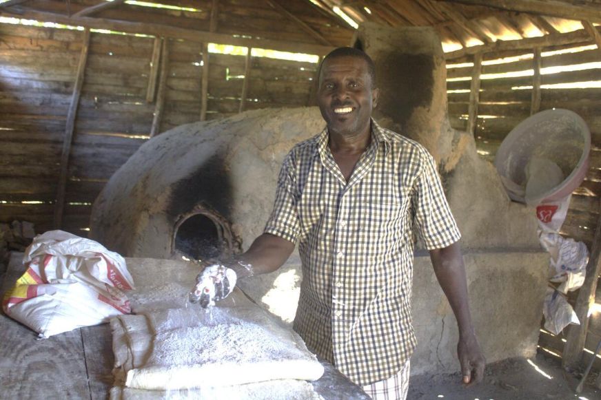 An adult stands inside a rustic bakery beside a large clay oven, holding flour and smiling. (Haiti, 2024)