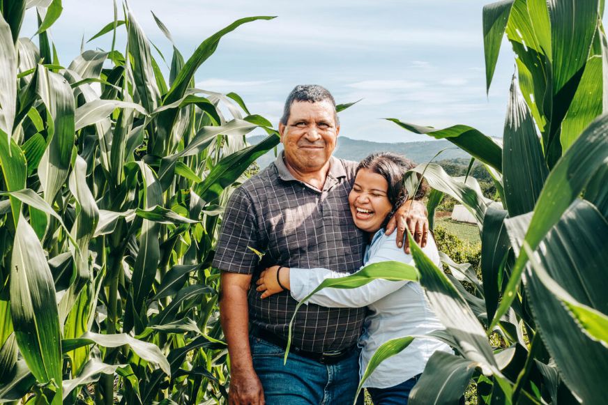An adult stands in a cornfield with a child holding onto them, surrounded by green plants in a rural landscape. (Honduras, 2025)