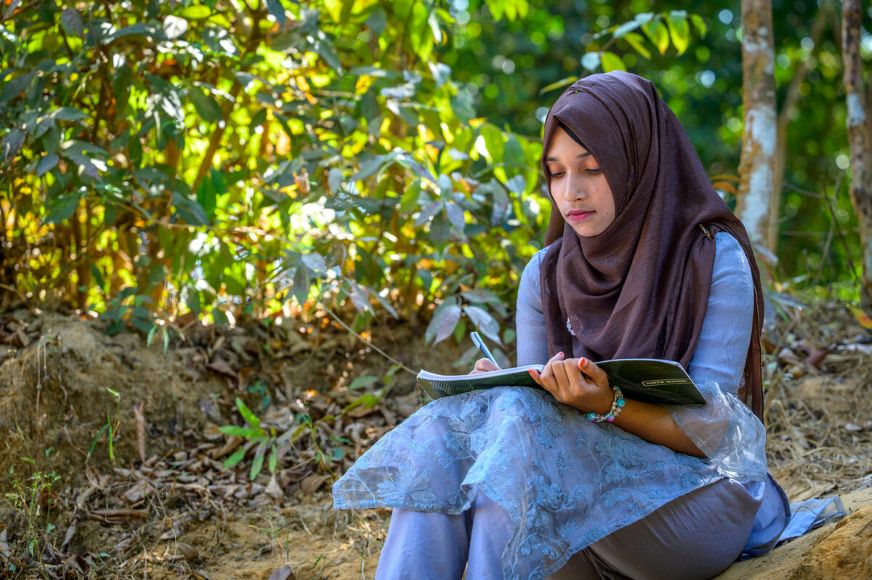 A young person sits outdoors on the ground near greenery, writing in a notebook, with trees and foliage in the background. (Bangladesh, 2025)