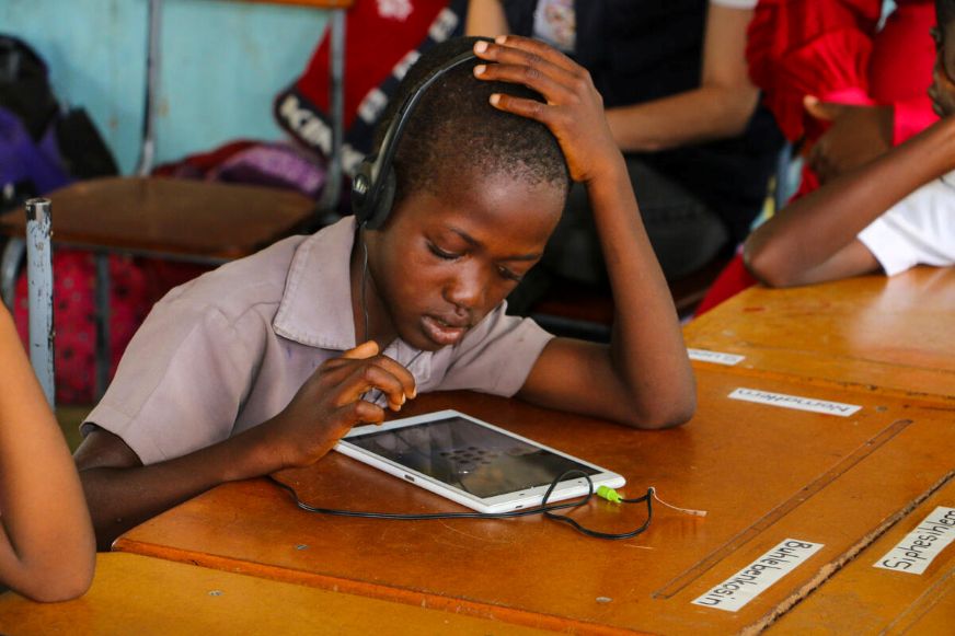 A child sits at a wooden desk wearing headphones, focusing on a tablet device with one hand on their head, in a classroom setting. (Zimbabwe, 2026)
