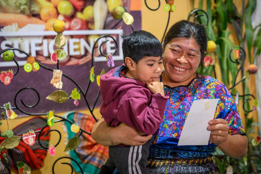 An adult holds a child while looking at a piece of paper together indoors, surrounded by colorful decorations and a display of fruits in the background. (Guatem