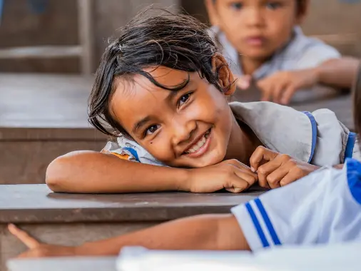 A young girl resting leaning on a desk while smiling at the camera.