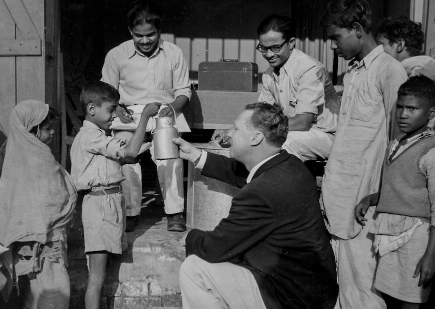 Sur une photo en noir et blanc, un garçon tient un pot à lait en métal à la main tandis qu’un homme vêtu d’une veste de costume noire lui parle.