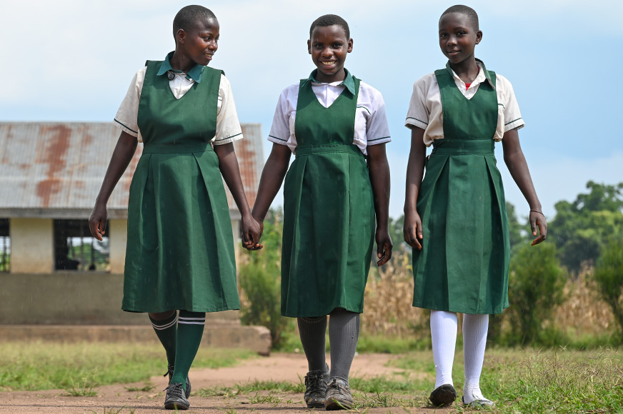 Trois adolescentes vêtues d’un uniforme scolaire vert marchent le long d’un chemin de terre dans un champ herbeux.