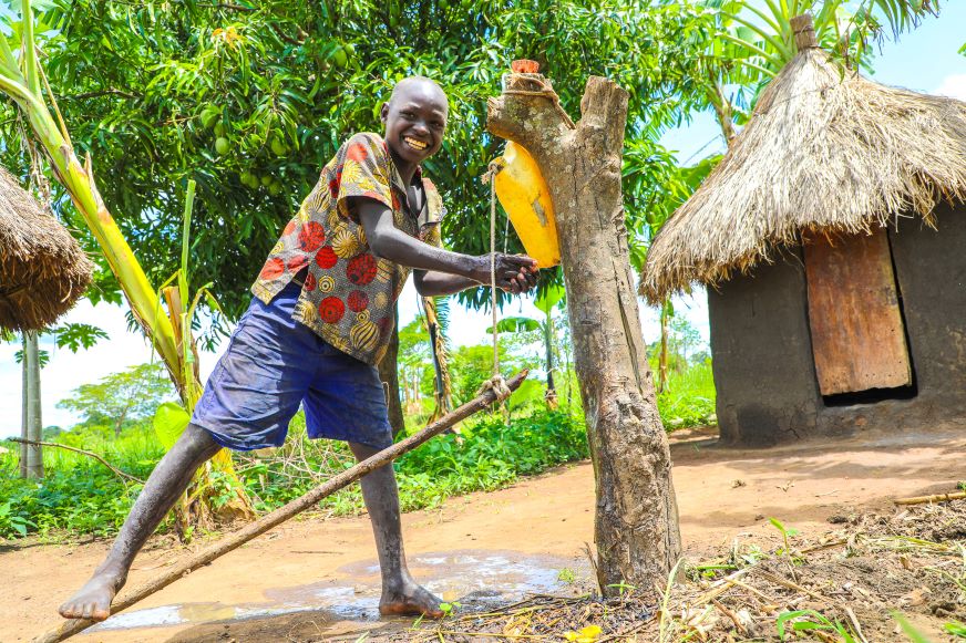 En Ouganda, un adolescent se lave les mains à l'aide d'un bidon d'eau jaune suspendu à une souche d'arbre.