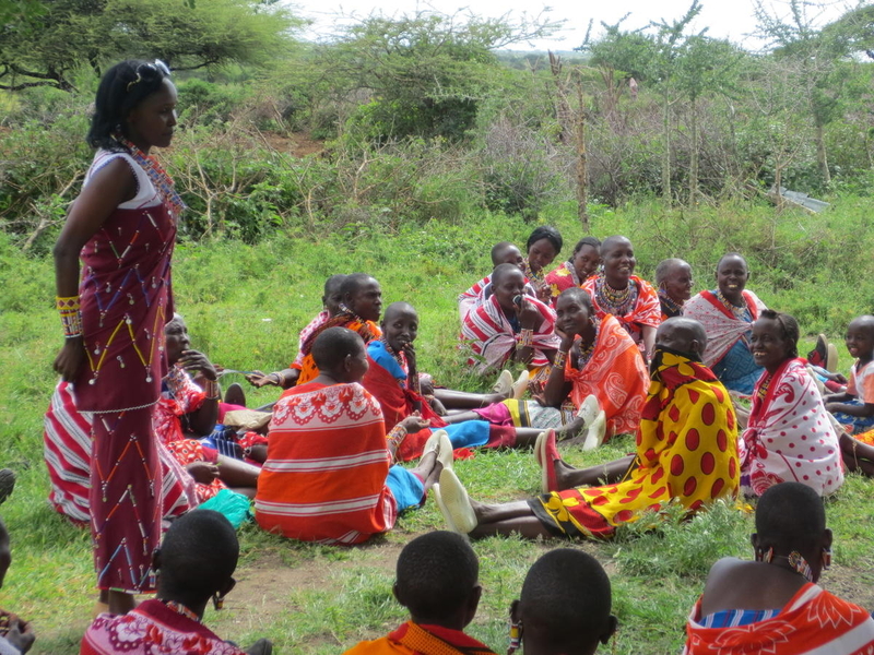 Un groupe de femmes vêtues de robes aux couleurs vives est assis sur l'herbe. Une femme se tient debout parmi elles.