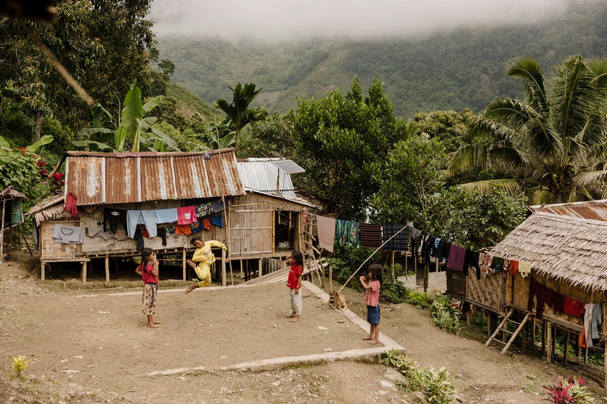Quatre enfants jouent dans une cour en terre battue, entourés de cabanes en bois sur pilotis et d’une végétation luxuriante, dans une région montagneuse. (Indonésie, 2023)
