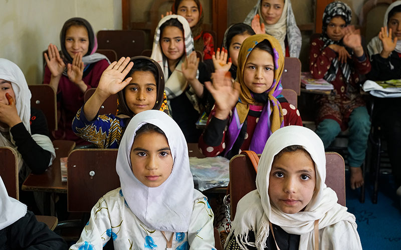 Dans une salle de classe en Afghanistan, des jeunes filles sont assises à leurs pupitres et lèvent la main.
