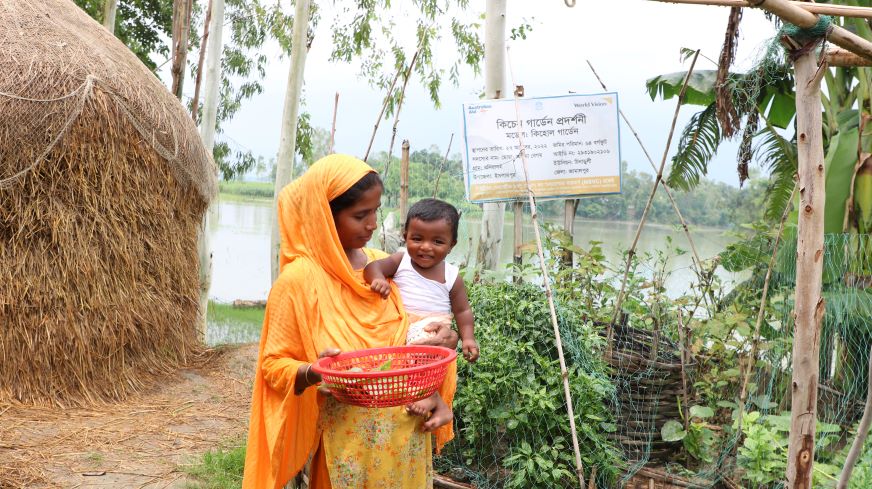 Au Bangladesh, une mère vêtue d’orange tient son bébé sur la hanche tout en cueillant des légumes dans son potager.