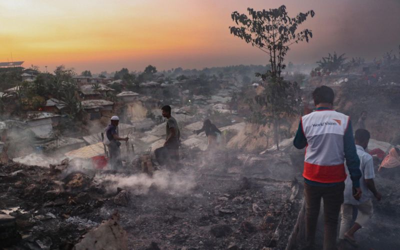 Un homme portant un chandail de World Vision se dirige vers les bâtiments d’un camp de réfugiés qui fument encore après un incendie.