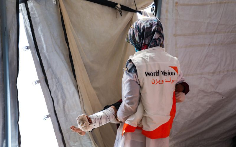 Un travailleur de World Vision sort d’une tente en portant un enfant dont la jambe est immobilisée par une attelle et bandée.