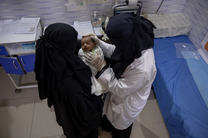 Deux femmes vêtues d’un niqab noir — dont l’une porte une blouse médicale blanche — tiennent un nourrisson entre elles.