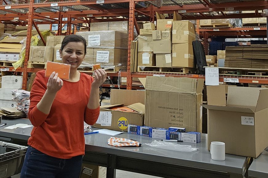 Une femme vêtue d'un chandail orange tient des fournitures d'emballage dans un entrepôt rempli de boîtes.