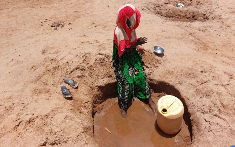 En Somalie, une femme est assise au bord d’un trou creusé dans le lit asséché d’une rivière. Ses pieds s’enfoncent dans la boue au fond du trou.