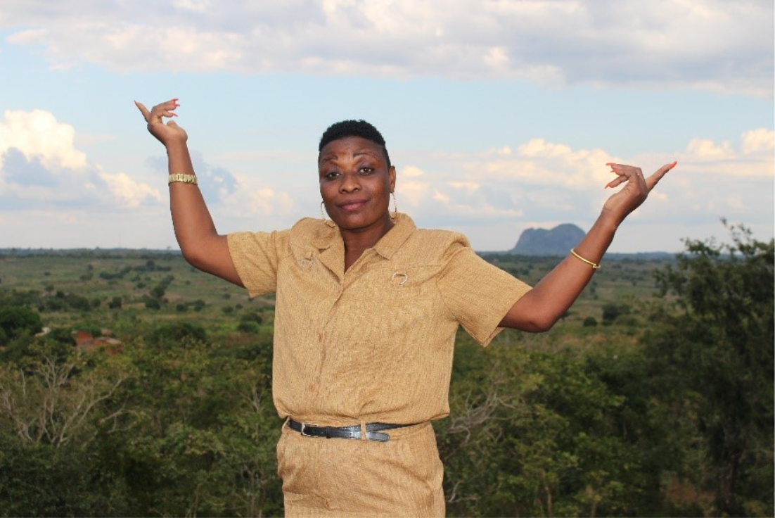 Une femme vêtue d’un uniforme brun clair se tient debout devant un paysage, les bras levés, dans un geste exprimant la fierté et l’accomplissement.