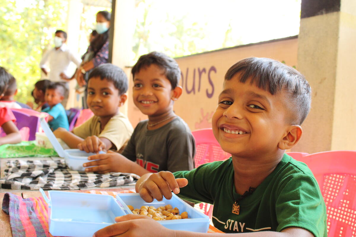 Des enfants sourient en dégustant leur dîner dans des contenants bleus, assis à une longue table.