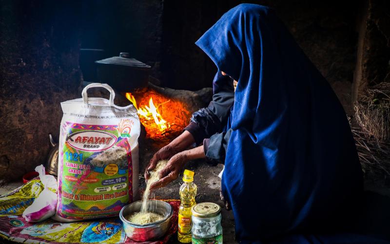 Une femme afghane, vêtue d’un hijab d’un bleu intense, s’accroupit sur un plancher de béton pour préparer du riz.