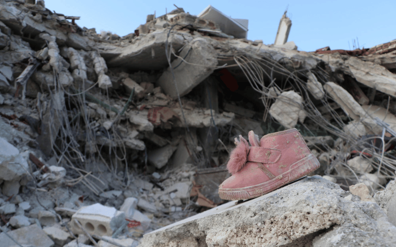 Une chaussure rose repose sur un morceau de béton brisé, au milieu des débris d’un immeuble en arrière-plan.
