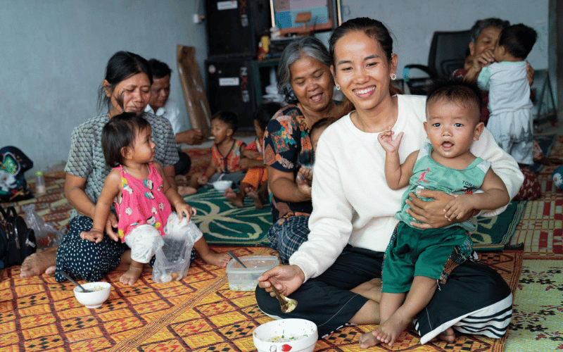 Un groupe de femmes cambodgiennes est assis sur des nattes, un bol de nourriture à la main et leur bébé sur les genoux.