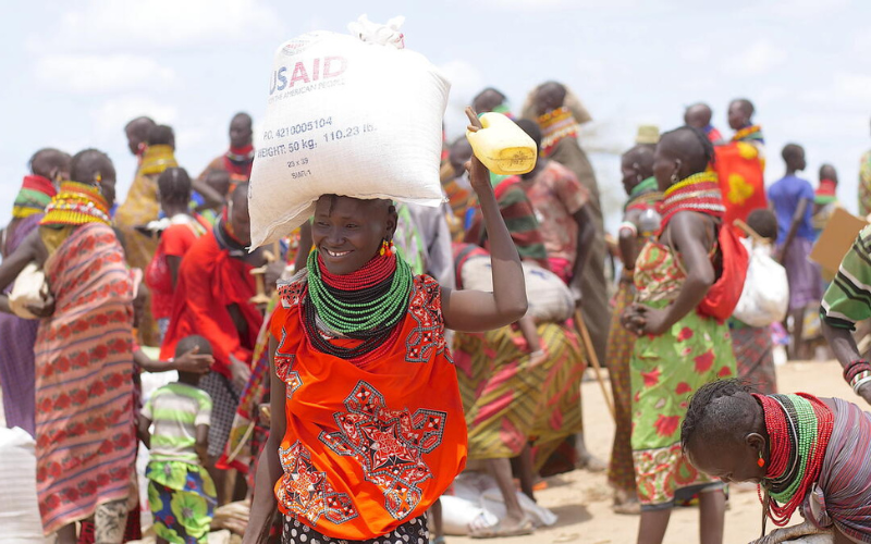 Une Kényane souriante porte un sac de provisions sur la tête, avec d’autres personnes en arrière-plan.
