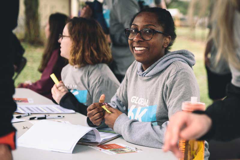 Une femme vêtue d’un chandail à capuchon gris sourit à la caméra. Elle est assise à une table avec d’autres bénévoles. Ils ont tous des documents devant eux.