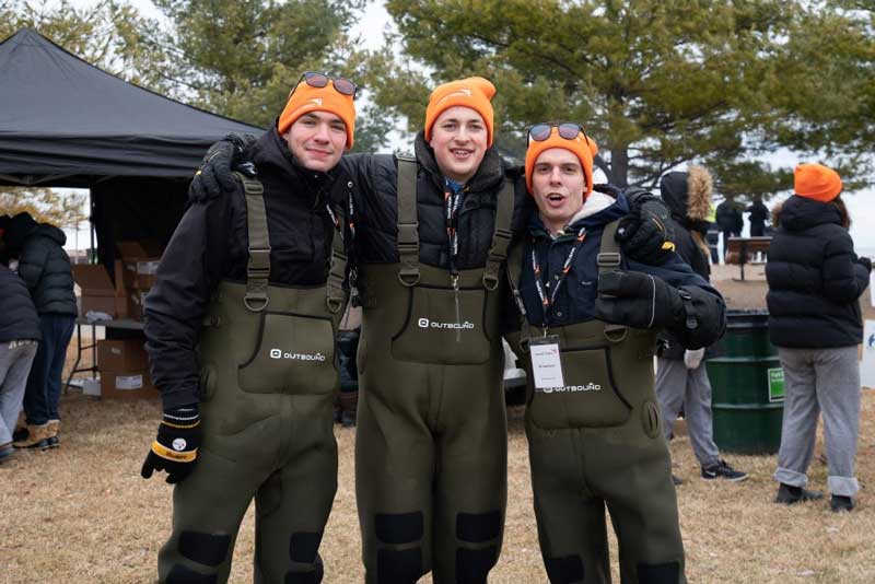 Trois hommes, coiffés de chapeaux orange, vêtus de manteaux noirs et de pantalons qui semblent imperméables, se tiennent par les épaules. Ils sourient à la caméra.
