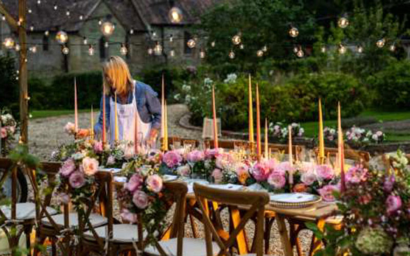 Une femme prépare une longue table pour un repas, à l'extérieur. La table est décorée de roses roses.