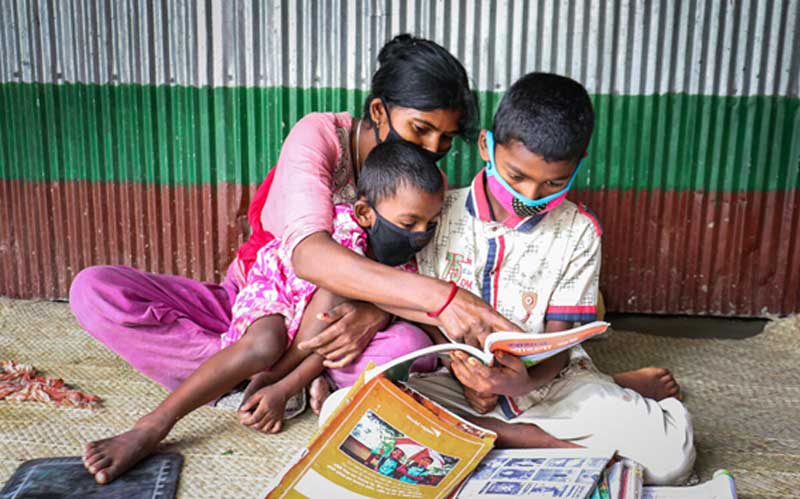 Une mère et ses deux enfants, tous munis de masques de protection, lisent des livres ensemble. Ils sont assis sur le plancher de leur maison.