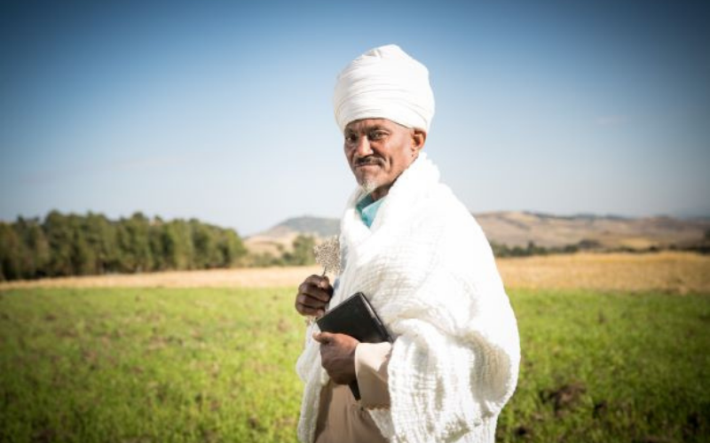 Un homme portant un turban blanc et une cape blanche sourit à la caméra.