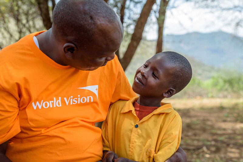 Un homme adulte, vêtu d’un chandail orange de World Vision, regarde avec tendresse une petite fille souriante à West Pokot, au Kenya.