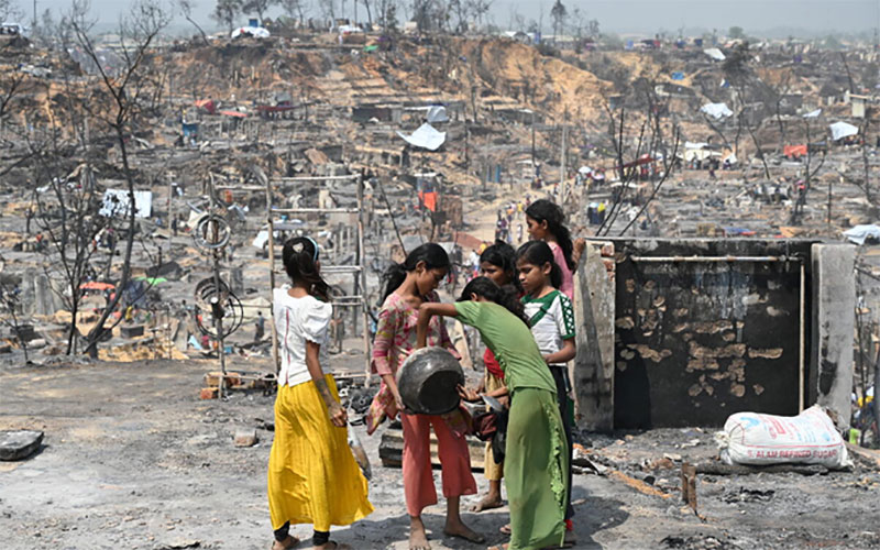 Dans le camp de réfugiés de Cox’s Bazar, un groupe de filles tient un bol ou une casserole et regarde à l’intérieur. Autour d’elles gisent les décombres calcinés du camp.