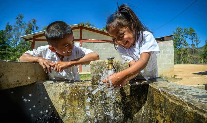 Au Honduras, une jeune fille en uniforme scolaire s’asperge les mains d’eau propre pour se laver. Elle arbore un large sourire tandis que son amie l’observe.