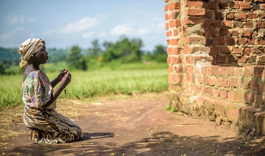 Une femme est agenouillée dans la terre devant un édifice en brique, une croix entre les mains.