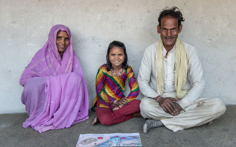 Une jeune fille indienne est assise par terre chez elle. Ses parents sont assis de chaque côté d’elle et ils sourient tous les trois. Il y a un livre devant eux.