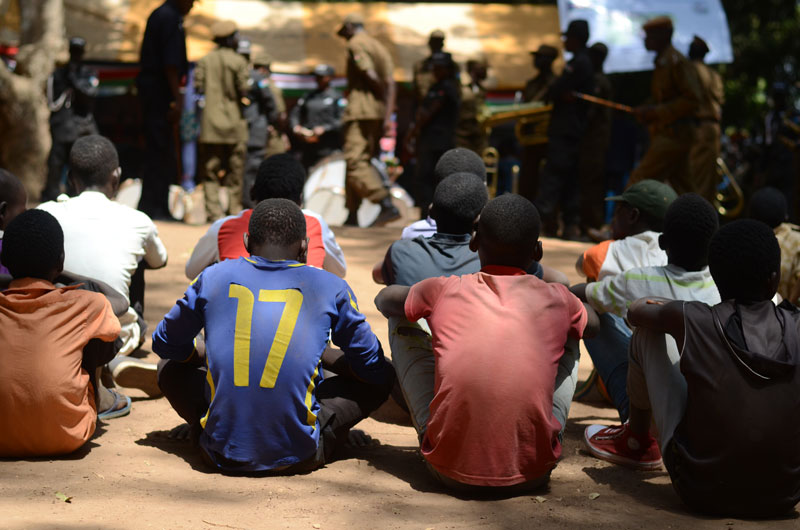 Des adolescents, anciens enfants soldats, sont assis sur un sol en terre battue, dos à la caméra, dans l'attente d'une aide.
