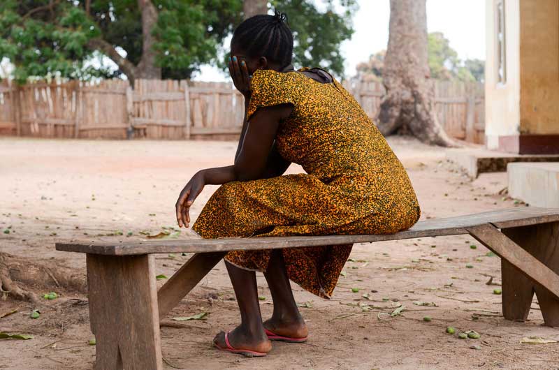 Une femme du Soudan du Sud, vêtue d’une robe jaune, est assise sur un banc de bois, dos à la caméra.