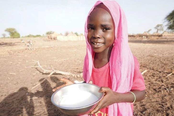 Au Soudan, une petite fille coiffée d’un foulard rose se tient debout au milieu d’un paysage aride, tenant dans ses mains un bol de lait de chèvre frais et blanc. Elle sourit.