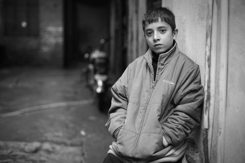 Un jeune garçon aux cheveux foncés est adossé à un mur de briques dans une ruelle. Il porte un manteau d’hiver trop grand pour lui et regarde droit vers la caméra.