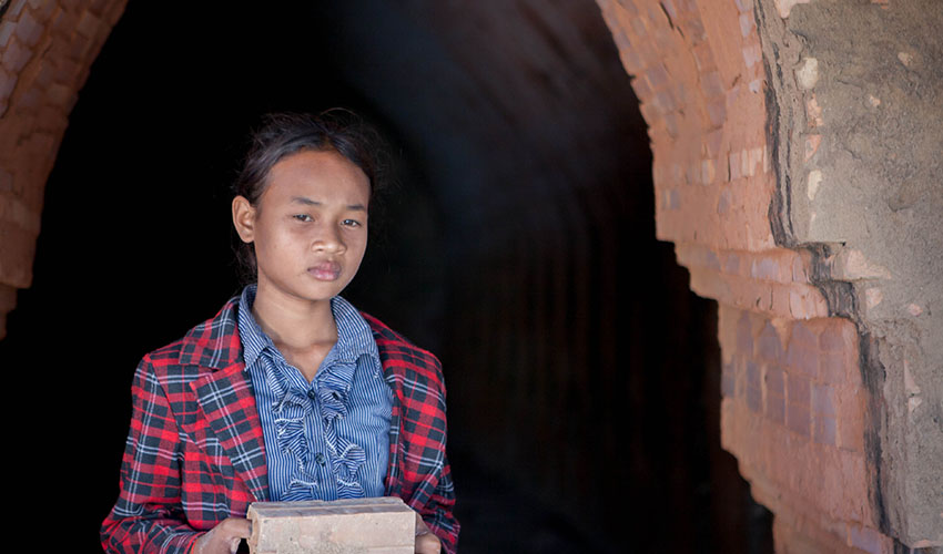 Une fillette cambodgienne tient quelques briques d'argile dans les mains et se tient dans l'embrasure d'une porte, l'air sombre.