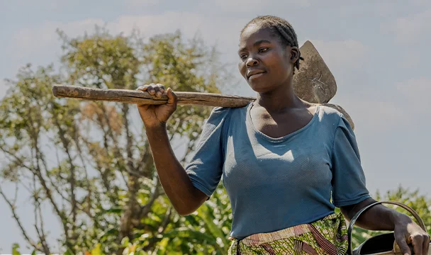A woman in a blue shirt carrying a hoe and watering can outdoors, surrounded by greenery.