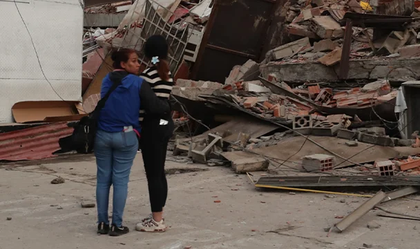 Two women stand near a large pile of rubble and collapsed concrete buildings in an urban area.
