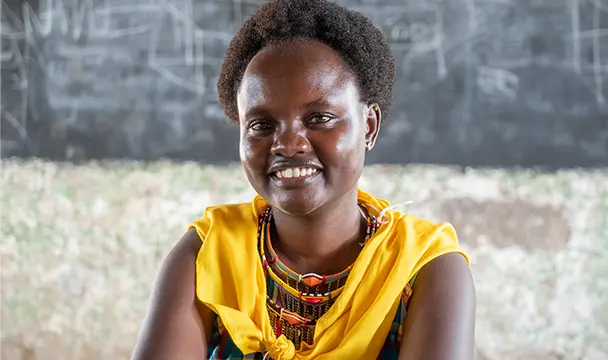 A young woman draped in a yellow scarf sits in front of a chalkboard covered with writing. (Kenya, 2018)