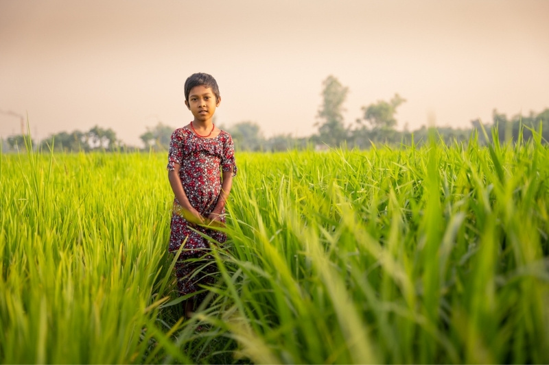 A child in Bangladesh smiles for a photo next to crops that were grown with the help of the REACTS-IN program.